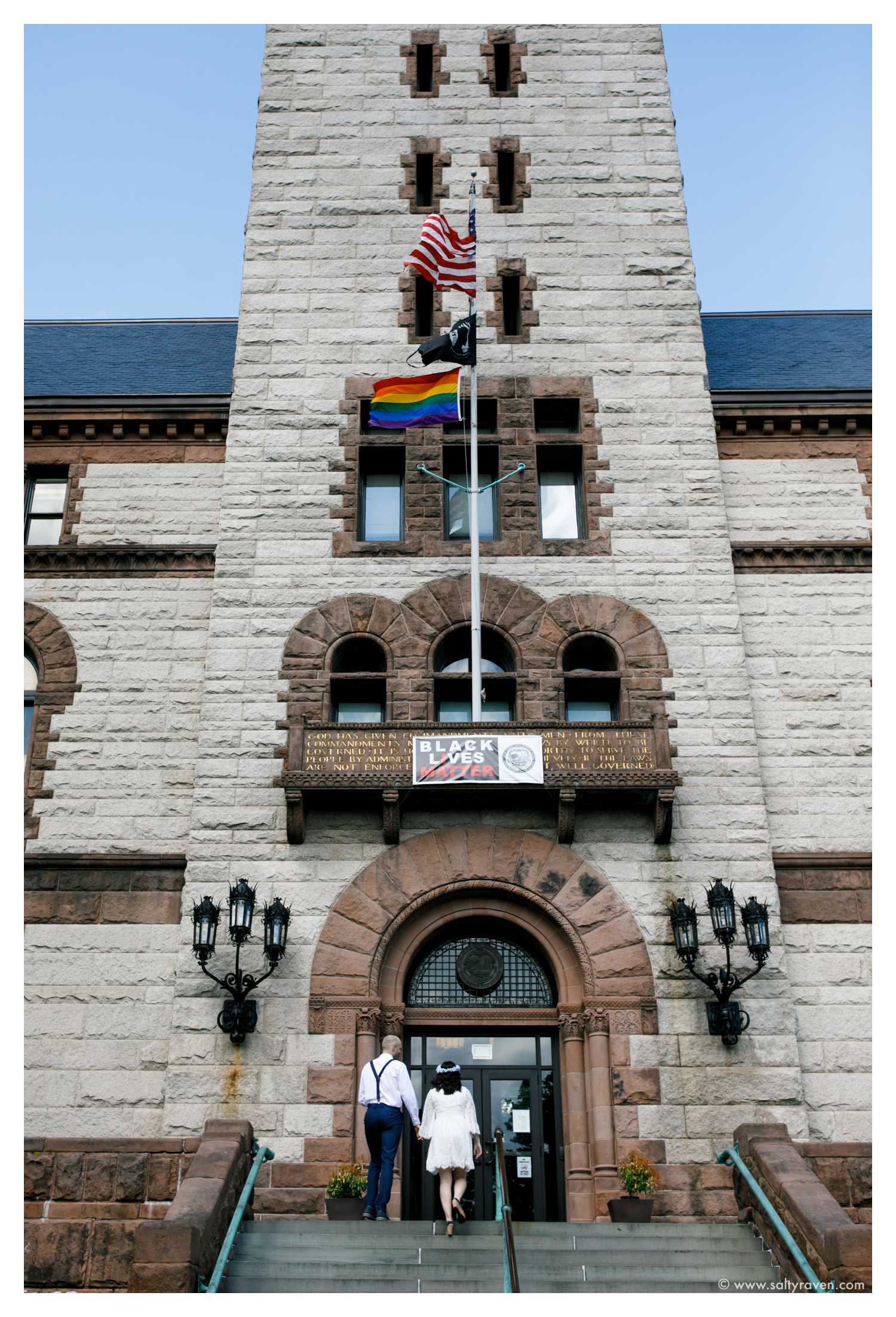 The couple walks up the steps to Cambridge City Hall. The American Flag flies along with others including the Gay Pride flag. 