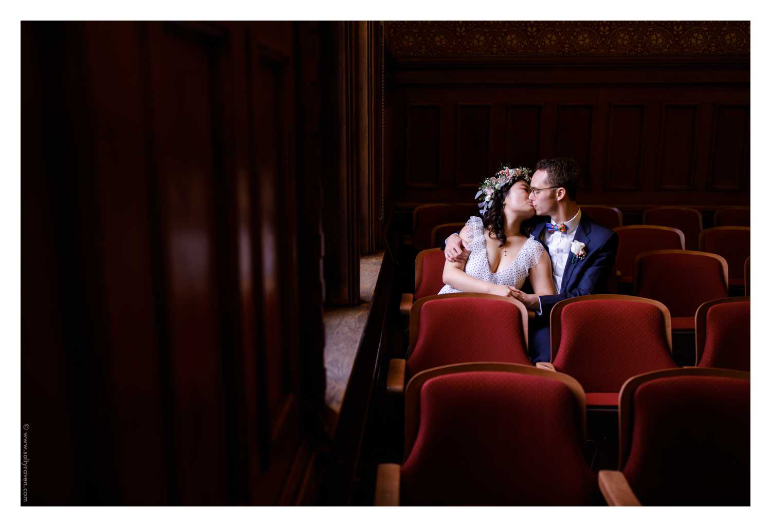 The couple steals a kiss in the red seats near a window at their Cambridge City Hall Wedding.